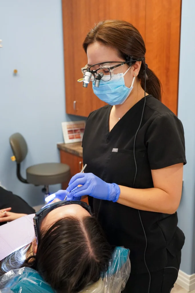 Female dentist Dr. Peggy Chuang wearing magnifying dental loupes, a surgical mask, and blue gloves performing a precision procedure on a patient.