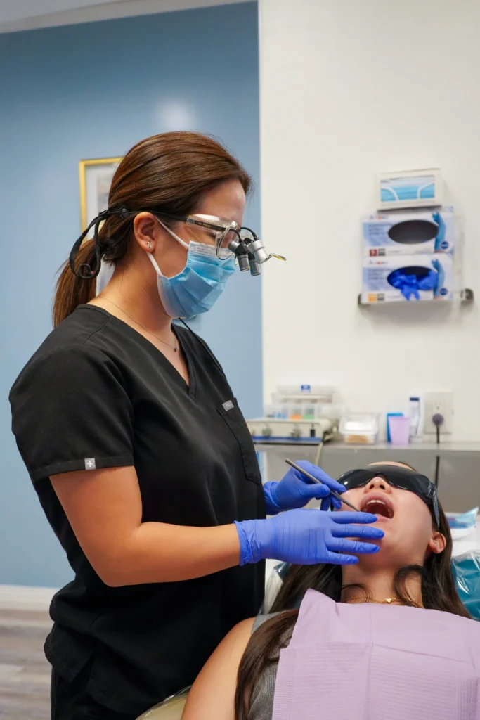 Profile shot of female dentist Dr. Peggy Chuang in black scrubs and a surgical mask examining a patient's open mouth with specialized dental instruments.