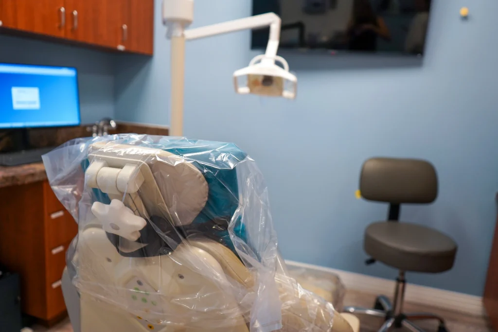 Clean, modern dental treatment room featuring an ergonomic patient chair with protective plastic covers, clinical stools, and digital screens against light blue walls.