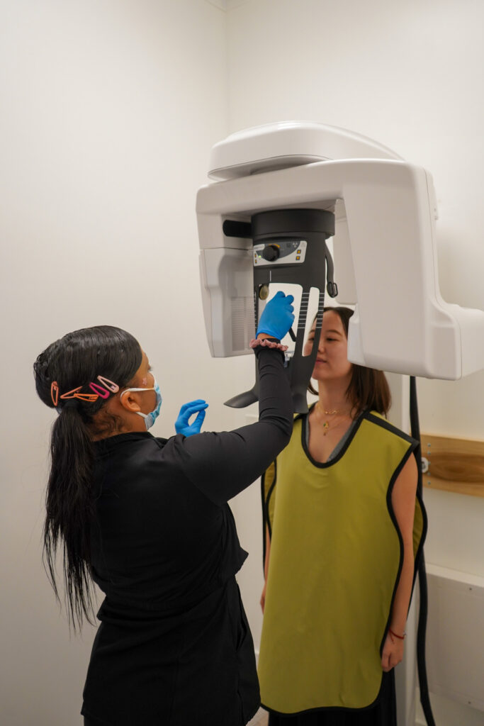 Dental assistant adjusting a modern panoramic CBCT X-ray machine for a standing female patient wearing a yellow protective lead apron.