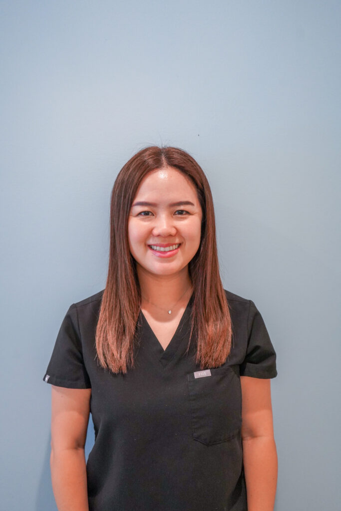 Professional headshot of female dentist Dr. Peggy Chuang smiling warmly in black scrubs against a soft blue background.