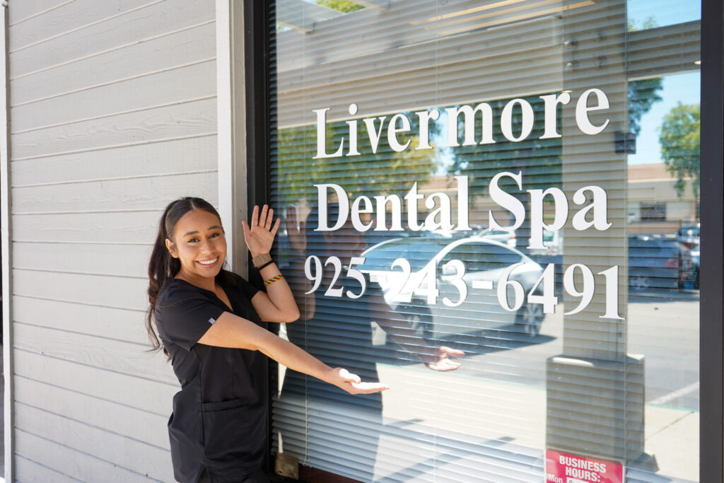 Smiling dental team member in black scrubs standing outside the clinic, pointing to the white Livermore Dental Spa window logo and phone number.